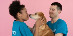 Two volunteers in matching blue t-shirts with an organization's name share a tender moment with a golden-colored dog, with the woman touching noses with the dog while the man smiles, all against a pink background