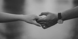 Black and white photograph of two hands reaching out to touch each other, one wearing a wristwatch, symbolizing connection and intimacy against a blurred background