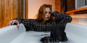 Young woman wearing a black and white plaid shirt lying in an empty white bathtub, with red hair and lipstick, looking pensively to the side with wooden paneled walls in the background