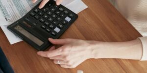 Close-up of hands using a black calculator with medical billing forms visible on wooden desk