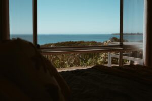 A serene coastal view seen through a large open window from a bedroom. The foreground shows a darkened bed with soft bedding, while the background reveals lush greenery leading to a calm blue ocean under a clear sky. Sunlight filters through, creating a peaceful and relaxing atmosphere.