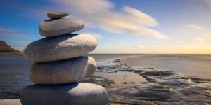 A carefully stacked pile of smooth, rounded stones sits in the foreground on a rocky shoreline. The background features a calm ocean stretching to the horizon under a soft blue sky with wispy clouds.