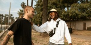 Two young men are seen laughing and enjoying a conversation in an outdoor park setting. One is wearing a black t-shirt and has a shaved head with earrings, while the other is wearing a white hoodie, a beige cap, and a crossbody bag.