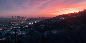 A breathtaking view of Los Angeles at sunset, with the sky displaying hues of pink, orange, and purple. The city skyline in the background is illuminated by the glow of streetlights and buildings, while the hills in the foreground are dotted with houses and winding roads.