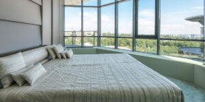 A modern bedroom with a large, comfortable bed covered in white linens, positioned against floor-to-ceiling windows that offer a breathtaking panoramic view of a green cityscape.