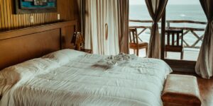 A beautifully arranged bedroom with a large, neatly made bed covered in white bedding, facing a wooden balcony overlooking the ocean.