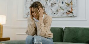 A young woman with light brown hair sits on a green couch, holding her head in her hands with a distressed expression.