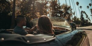 A man and a woman cruise through a palm tree-lined street in a vintage convertible under the warm afternoon sun. The driver, wearing sunglasses and a dark shirt, grips the wheel while the passenger, a woman with long blonde hair, looks ahead.