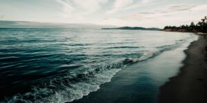 A peaceful beach scene with gentle waves washing onto the shore under a cloudy sky, with distant cliffs and trees lining the horizon.