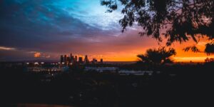 A panoramic view of the Los Angeles skyline at sunset, with high-rise buildings silhouetted against a dramatic sky of orange, pink, and blue hues.
