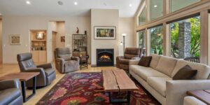A cozy and stylish living room featuring a modern fireplace, comfortable seating, a colorful floral rug, and large windows offering a scenic view of a lush garden and stone columns