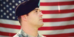 A close-up of a solemn U.S. soldier in uniform and beret, looking upward with the American flag softly blurred in the background.