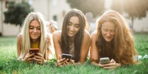 Three teenage girls lying on grass and smiling while using their smartphones, highlighting the everyday role of social media in youth connection, communication, and digital culture.