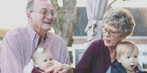 An elderly couple sitting outdoors and smiling while holding a baby and a toddler, enjoying a happy moment together with their grandchildren.