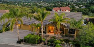 Aerial view of a well-maintained single-story house with palm trees, a landscaped front yard, and decorative lighting, set against a backdrop of rolling hills and a suburban neighborhood.