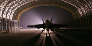 A silhouetted fighter jet parked inside a large, illuminated hangar at night, with shadows stretching across the runway under a bright backlight.
