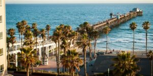 A scenic coastal view of Oceanside Pier extending into the blue Pacific Ocean. Tall palm trees line the beach promenade, with a sandy shore and beachgoers visible below. A white building appears on the left side, and the wooden pier structure stretches far into the water against a clear sky. The scene is bathed in warm late afternoon light.