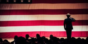 A uniformed military officer salutes on stage in front of a large American flag, with a group of individuals silhouetted in the foreground during a patriotic or military event.