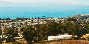 A panoramic view from a hillside overlooking a coastal city with residential neighborhoods, dense trees, a long pier extending into the ocean, and distant mountains across the water.