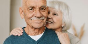 An elderly man smiles warmly at the camera while a joyful elderly woman hugs him from behind, her face lit up with laughter and affection.