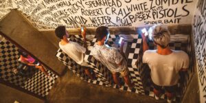In a quirky, graffiti-covered restroom, young people stand at urinals and sit in stalls while fully absorbed in their smartphones—highlighting the ever-present pull of social media, even in private spaces.