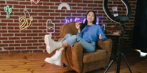 A young woman sits in a cozy room with neon signs and a pizza slice, recording content for social media with a smartphone and ring light—capturing the modern lifestyle of digital influencers.