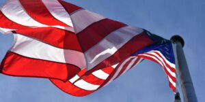 A close-up view of the U.S. flag rippling in the wind atop a flagpole, with vibrant red, white, and blue colors standing out sharply against a bright blue sky symbolizing freedom and national pride.