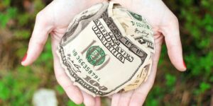 A person holds a crumpled ball made entirely of U.S. hundred-dollar bills, symbolizing financial decisions, economic value, or the impact of money in human hands.