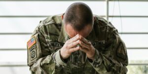 A U.S. Army soldier sits with his head bowed and hands clasped in a moment of deep reflection, symbolizing the emotional toll and inner strength associated with military service.