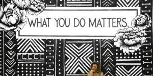 A woman sits beneath a bold, black-and-white mural that reads “What You Do Matters,” surrounded by floral art and geometric patterns, an inspiring visual reminder of purpose and impact.
