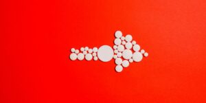 A collection of white circular pills arranged in the shape of a right-facing arrow on a bright red background.