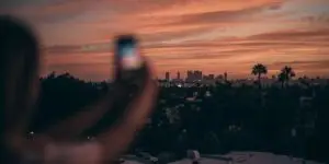 scenic view of a city skyline and palm trees silhouetted against a pink and orange sunset, with a blurred hand holding a smartphone in the foreground capturing the scene.