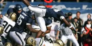 An American football player in a blue jersey is captured mid-air, flipping upside down over a pile of opposing players in white and gold helmets during a crowded game.