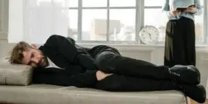 Man struggling with depression lying on couch during addiction recovery while partner maintains emotional distance, illustrating challenges of dual diagnosis treatment