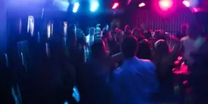 A motion-blurred photograph of a crowd of people dancing in a dark nightclub, illuminated by bright blue and pink stage lights.