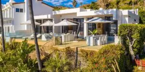 A beautiful, multi-level luxury rehabilitation home featuring a spacious sun deck with white umbrellas, outdoor seating, and glass railings overlooking the scenic coastal landscape.