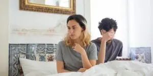 A distressed man and woman sitting apart on a bed in an upscale, brightly lit room, illustrating the emotional toll of substance use disorders on relationships and the specialized counseling provided in private couples rehabilitation programs.