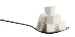 A close-up view of a silver spoon overflowing with stacked white sugar cubes against a plain white background, illustrating the concept of sugar addiction, dietary habits, and the risk of substituting substances with sweets during recovery.