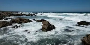 A wide, scenic view of a rugged coastline with white ocean waves crashing against dark rocks under a clear blue sky, illustrating the calming environment and therapeutic benefits of ocean therapy in a luxury addiction recovery program.