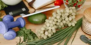 A close-up view of a wooden table arranged with fresh green grapes, an avocado, leafy herbs, a digital scale, and two blue dumbbells, illustrating the vital combination of a nutrient-rich diet and regular physical exercise for healing liver damage during alcohol addiction recovery.