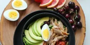 A top-down view of a nutrient-rich, balanced meal featuring a dark bowl of lean chicken, sliced avocado, and a hard-boiled egg, accompanied by fresh cherries, grapefruit slices, and tomatoes on a rustic wooden board, illustrating the critical role of a healthy diet in stabilizing mood, repairing brain chemistry, and reducing relapse risk during addiction recovery.
