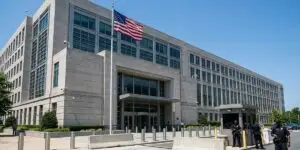 A large federal government building with an American flag on a flagpole, concrete security bollards at the entrance, and uniformed security officers standing at the front gate under a clear blue sky.
