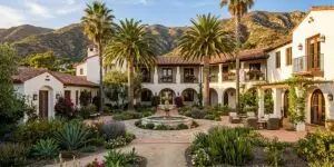 A Spanish Colonial-style treatment facility in California with white stucco walls, terracotta roof tiles, arched doorways, palm trees, a stone fountain in a landscaped courtyard, and green mountains in the background.