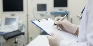 A close-up of a gloved hand writing on a clipboard with lined paper in a clinical setting, with medical monitoring equipment and screens visible in the blurred background.