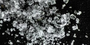 A macro photograph showing clear, jagged, and translucent crystals scattered on a dark black surface, representing crystal methamphetamine.