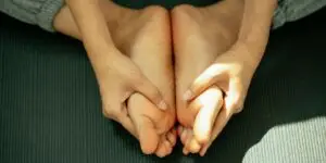 A close-up view of a person's hands gently holding their bare feet together on a dark yoga mat, illustrating grounding techniques, physical mindfulness, and the release of stored trauma practiced during somatic therapy in addiction rehabilitation.