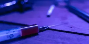 A close-up view of a syringe resting on a wooden surface under harsh dark blue lighting, illustrating the severe reality of intravenous drug use and the physical manifestation of how severe substance addiction compulsively alters and rewires brain chemistry.